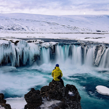 A man sits near a frozen waterfall, surrounded by icy terrain, showcasing the beauty of extreme geographical tourism.